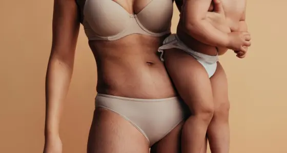 Close-up of a woman’s midriff after a mummy makeover holding her baby against a beige background