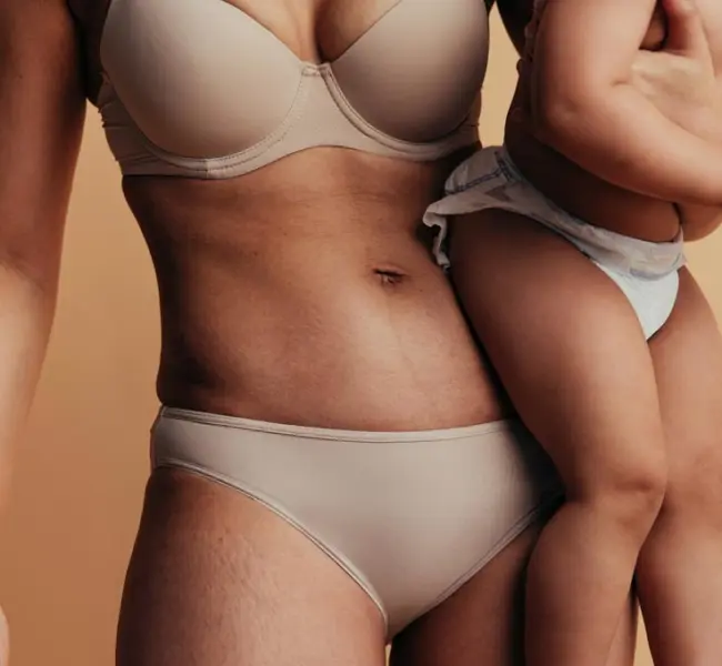 Close-up of a woman’s midriff after a mummy makeover holding her baby against a beige background