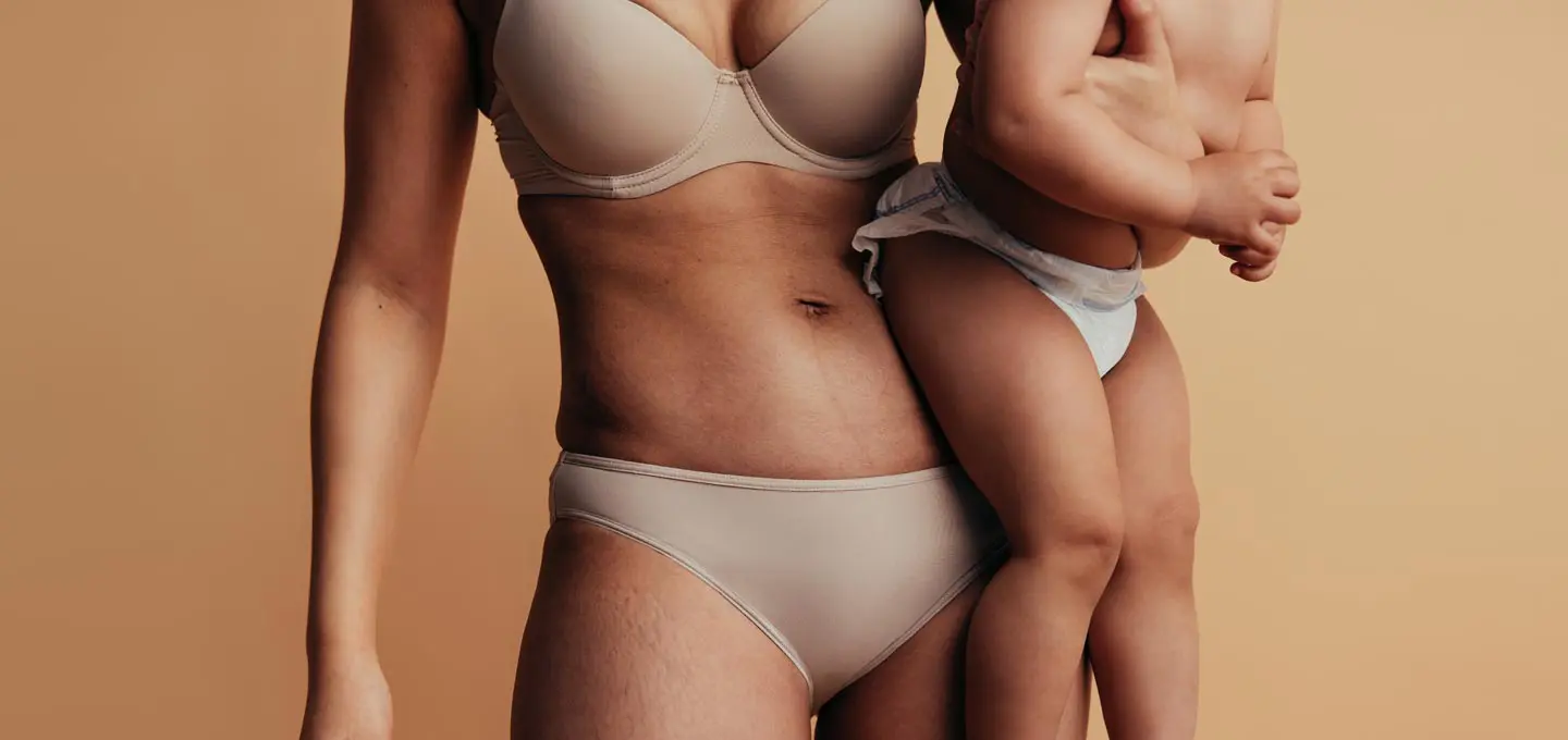 Close-up of a woman’s midriff after a mummy makeover holding her baby against a beige background
